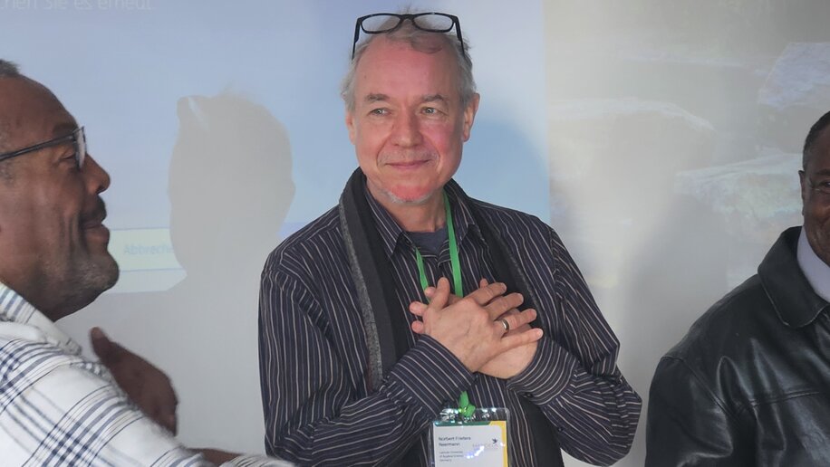 Drei Männer stehen vor einer Projektion mit dem Dell-Logo. Der Mann in der Mitte trägt einen Schal und ein Namensschild, er lächelt und hält die Hände vor der Brust. Die anderen beiden Männer stehen links und rechts von ihm und unterhalten sich. Die Atmosphäre wirkt entspannt und freundlich.