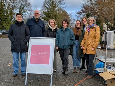 Eine Gruppe steht auf einem Schulhof zusammen und blickt in die Kamera. Im Vordergrund steht ein Aufsteller mit der Aufschrift "Kaffee ohne Filter".