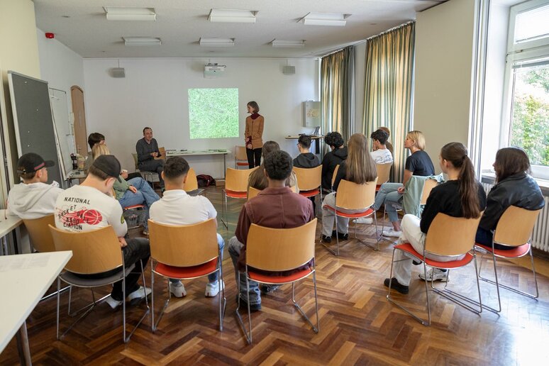 Junge Menschen sitzen in einen Raum und hören einen Vortrag zu. Dieser wird von einer Person gehalten, die vor der Gruppe steht. Auf der linken Person sitzt eine andere Person due mit der Gruppe kommunizeirt