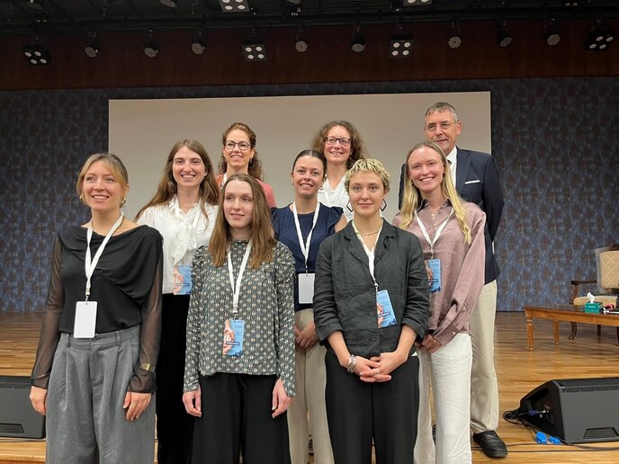 Eine Gruppe von neun Personen steht auf einer Bühne und lächelt in die Kamera. Die meisten tragen Namensschilder, was auf ihre Teilnahme an einer Veranstaltung hindeutet. Im Hintergrund ist eine Leinwand zu sehen.
