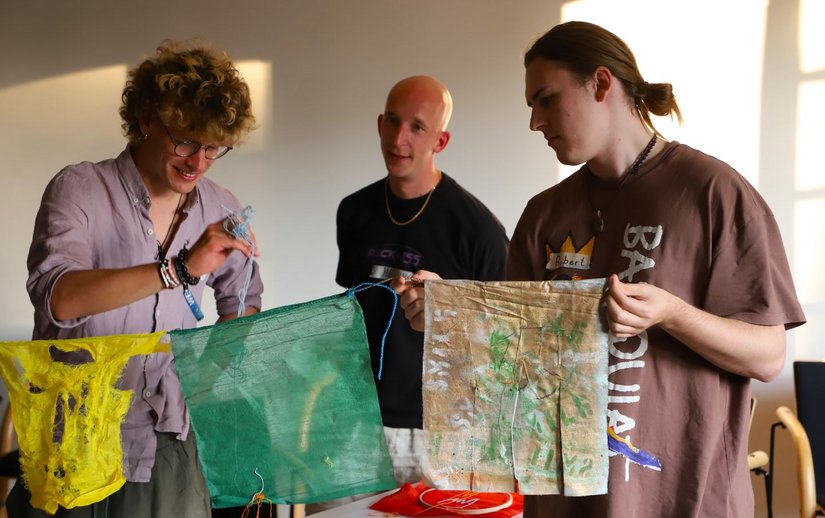 Drei Studierende in einem Raum: Einer hält eine gelbe, ein egrüne und eine beige bemalte Flagge an einer Schnur, ein anderer hält weitere Schnüre, der dritte beobachtet.die einer