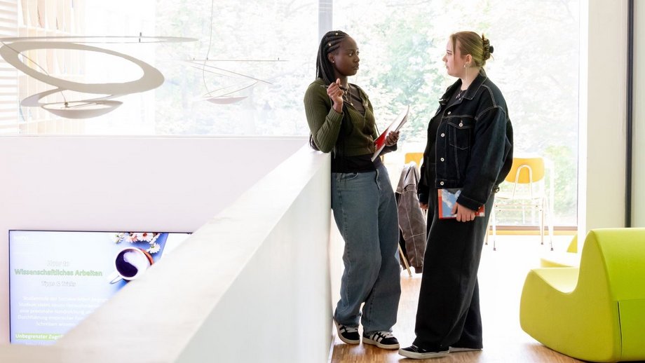 Zwei Studentinnen stehen in einem hellen Raum, halten Dokument in der Hand und sprechen miteinander. Der Raum hat ein große Fensterfront und ist mit modernen Hängelampen und grünen Sitzmöbeln ausgestattet.