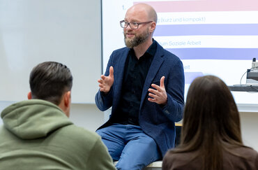 Ein Dozent mit Brille, Bart und dunkelblauem Jackett sitzt locker auf seinem Pult und erklärt etwas mit gestikulierenden Händen. Vor ihm sitzen zwei Studierende.