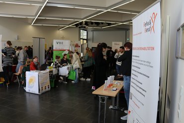 Das Bild zeigt einen Raum mit Aussteller_innen an ihren Ständen und Studierende, die diese Stände besuchen.