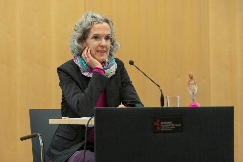 Eine Frau sitzt am Rednerpult. Sie hat Schulter lange graue Locken und trägt einen auffälligen bunten Schal und ein pinkes Oberteil.