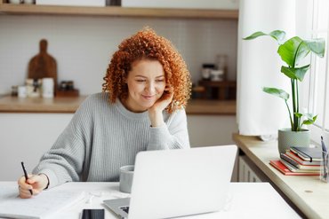 Eine Frau mit roten Locken schaut sich ein Online-Tutorial oder eine Lektion auf dem Laptop an, lächelt, hört zu und schaut aufmerksam, macht sich Notizen auf einem Block, sitzt am Küchentisch mit Smartphone und einer Tasse Kaffee.