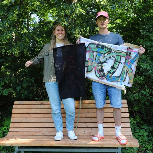Eine Studentin und eine Studentin stehen auf einer Bank und halten selbstgemalte Flaggen: Eine schwarze Flagge mit dem Wort "Macht", eine weiße Flagge mit grün-blauen Buchstaben und em Wort "Love".en.