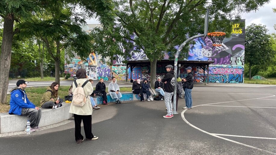 Auf einem von Bäumen umgebenen Basketballplatz steht eine kleine Gruppe von Menschen verteilt; einige unterhalten sich, andere schauen zu einem offenen Stand mit bunten Bildern im Hintergrund.