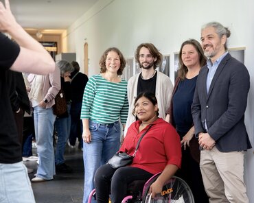 Die fünf Organisator_innen/Protagonist_innen/Künstler_innen (drei Frauen, zwei Männer) stehen nebeneinander vor den Fotografien und lächeln in die Kamera.