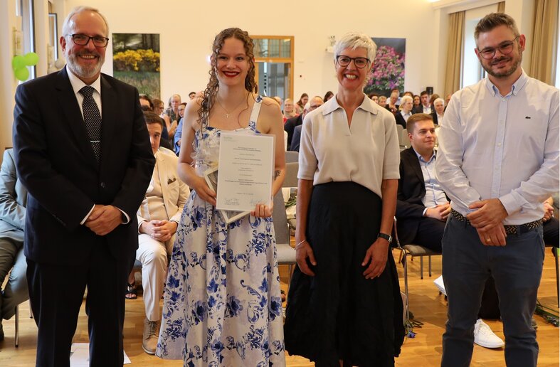 Eine Frau hält eine Urkunde und ein Geschenk in der Hand, neben ihr blicken zwei Männer und eine Frau in die Kamera.