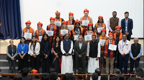Gruppenfoto auf einer Bühne bei einer Zertifikatsverleihung. Etwa 25 Personen stehen in zwei Reihen; in der hinteren Reihe tragen mehrere Personen orangefarbene Schärpen und rote Kopfbedeckungen und halten Urkunden.