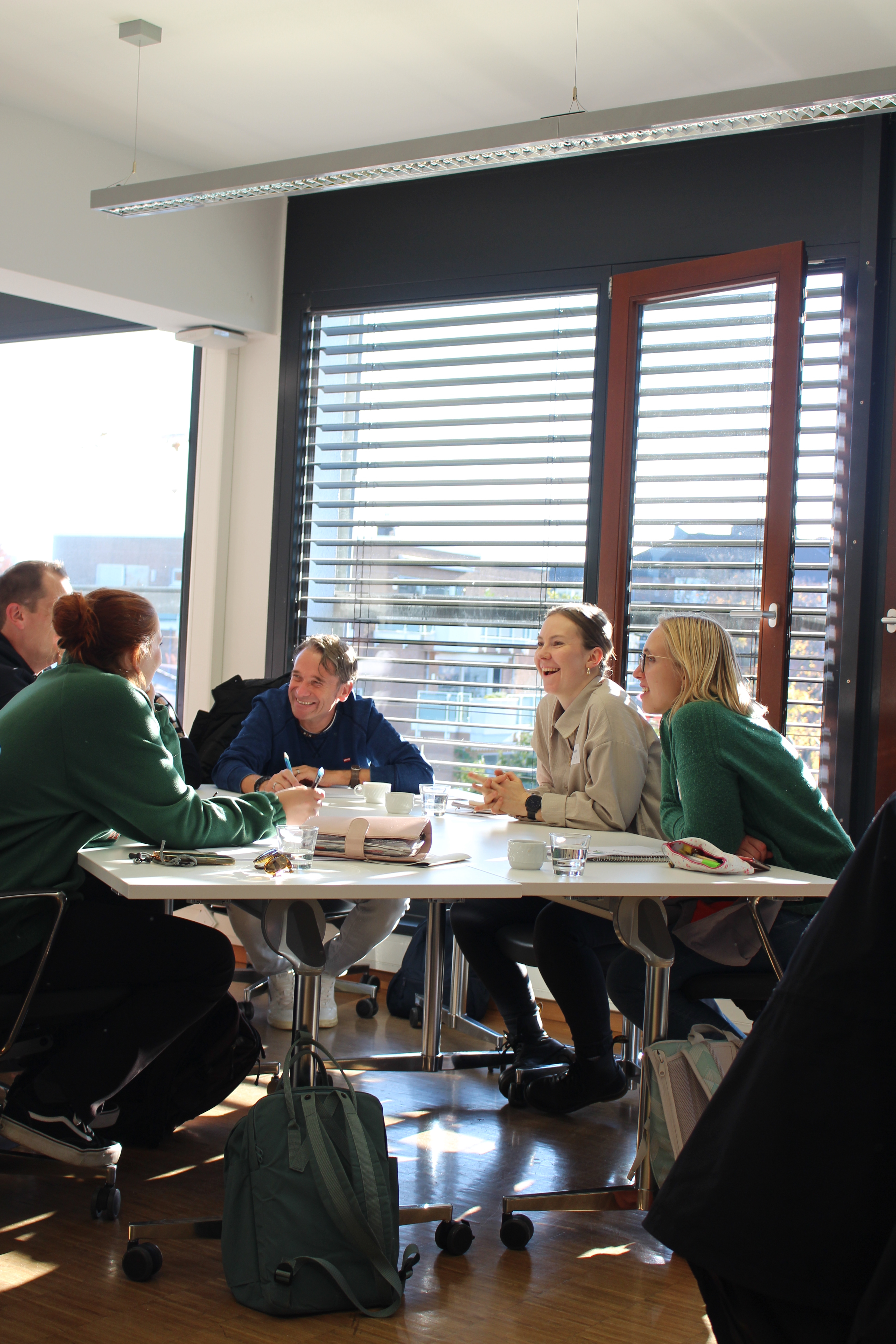 Sechs Erwachsene sitzen in einem hellen Seminarraum um einen Tisch, sprechen und lachen; auf dem Tisch liegen Notizbücher und Tassen, Sonnenlicht fällt durch Jalousien.