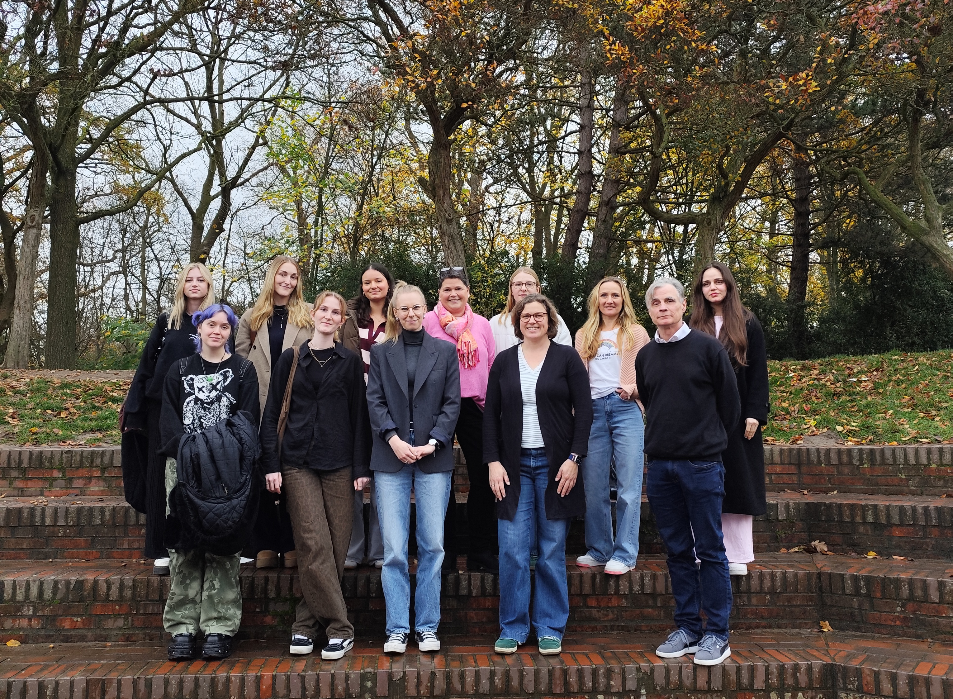 Eine Gruppe von Menschen, die vor einer Treppe aufgereiht stehen und freundlich in die Kamera blicken. Im Hintergrund befinden sich winterliche Bäume.