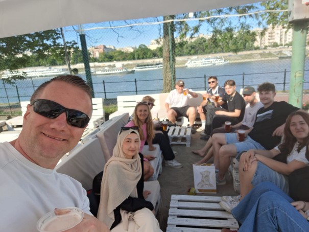 Eine Gruppe junger Personen sitzt unter einem Sonnensegel direkt an der Donau.