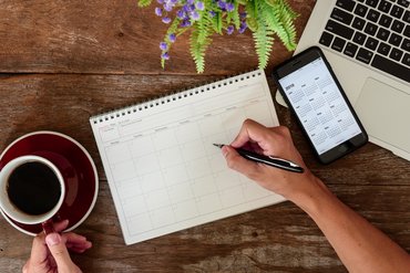 Nahaufnahme auf die Hand einer Person, die mit einem Stift etwas in einem Kalender notieren möchte; daneben liegt ein Organizer auf einem Laptop, die andere Hand hält eine Tasse Kaffee fest