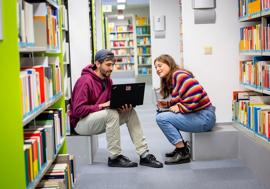 Zwei Studierende sitzen in der Bibliothek zwischen den Bücherregalen und schauen gemeinsam auf einen Laptop.