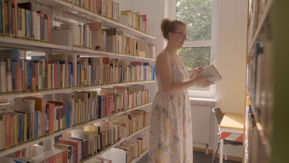 Eine junge Studierende steht in der Bibliothek und schlägt ein Buch in ihrer Hand auf.