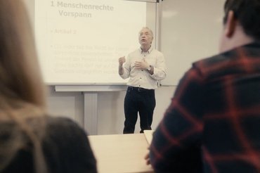 Ein Professor steht vor einer Präsentation und spricht zu Studierenden.