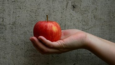 Eine Hand hält einen roten Apfel vor einem grauen Hintergrund.