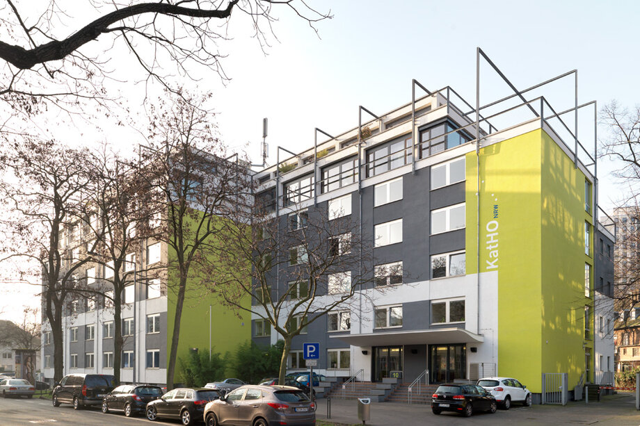 ein großes grau grünes Gebäude an einer befahrenen Straße. Auf dem grünen Teil des Gebäudes steht in großen Buchstaben "KatHO NRW".