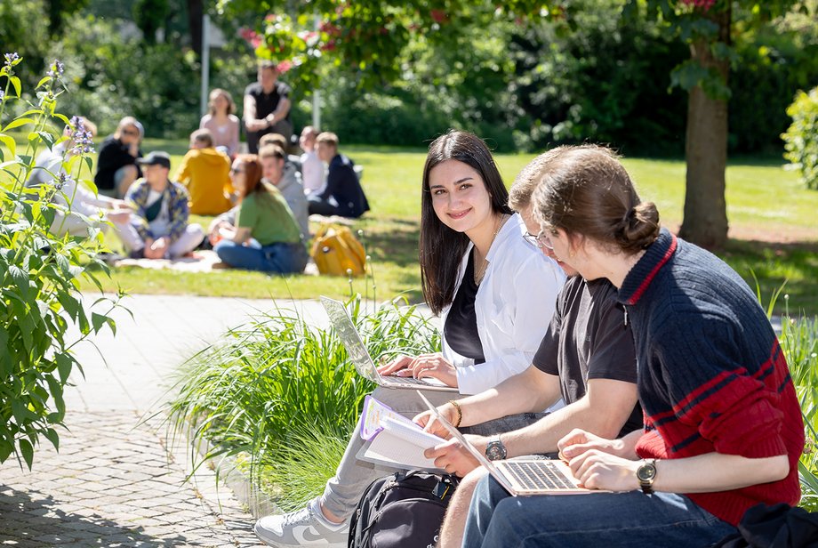 Mehrere Studierende halten sich im Außengelände auf. Drei sitzen auf einer Bank und unterhalten sich.

