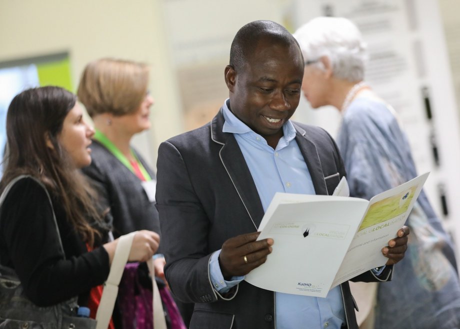 Ein Besucher des Social Glocalisation International Congress 2019 liest in der Broschüre zur Veranstaltung und lächelt dabei. Im Hintergrund unterhalten sich Besucherinnen miteinander.