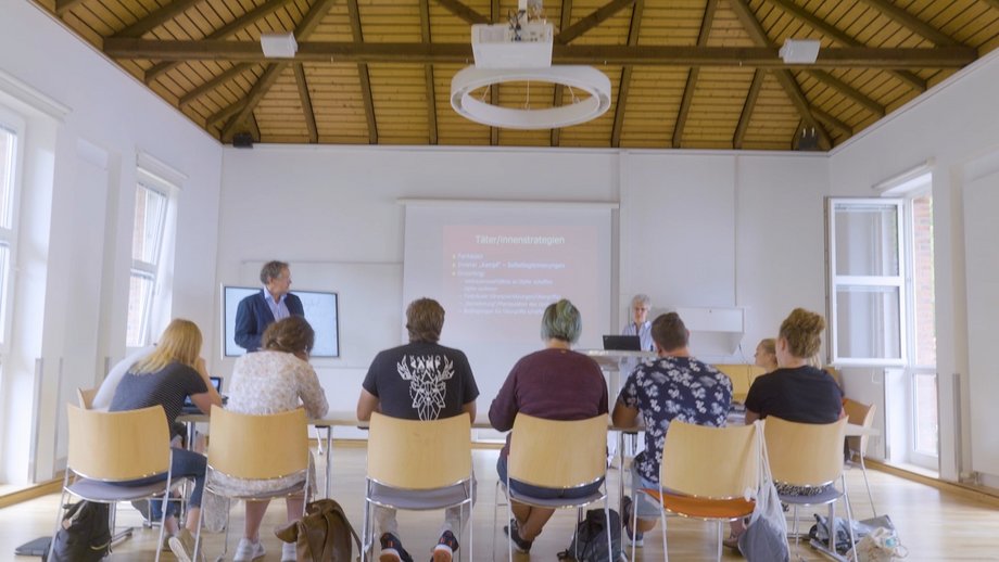 Eine Professorin hält in einem Seminarraum vor einer Gruppe Studierender eine PowerPoint-Präsentation.