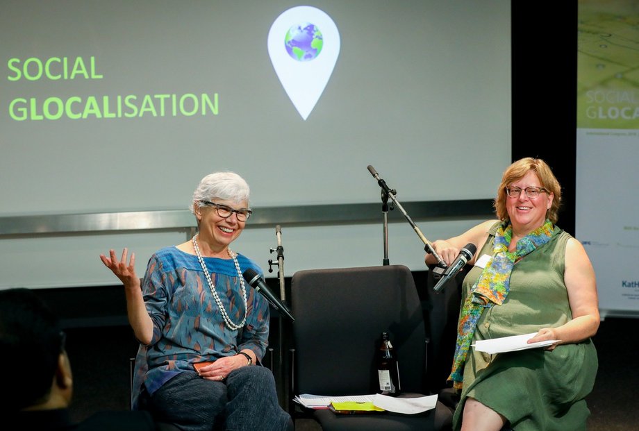 Zwei Frauen sitzen auf Stühlen vor Mikrofonen und beantworten Fragen während eines Symposiums. Sie lachen beide. Im Hintergrund ist eine Powerpoint-Präsentation mit der Aufschrift "Social Glocalisation" zu sehen.