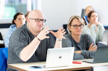 Mehrere Studierende sitzen in einem Seminarraum und hören einem Studenten zu.