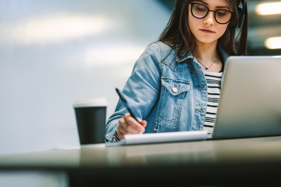 Eine junge Studierende sitzt mit Kopfhörern auf vor einem Laptop an einem Tisch und notiert sich etwas in einen Schreibblock.