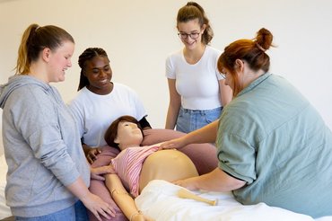 Mehrere Frauen stehen um ein Bett in dem eine schwangere Frauenpuppe liegt und üben den Umgang mit Schwangeren.