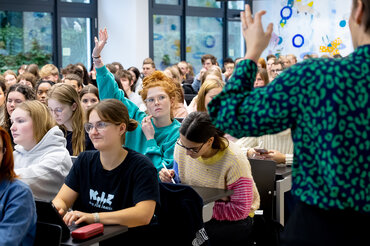 Mehrere Studierende sitzen in einem Seminar zusammen.
Eine Studentin meldet sich.