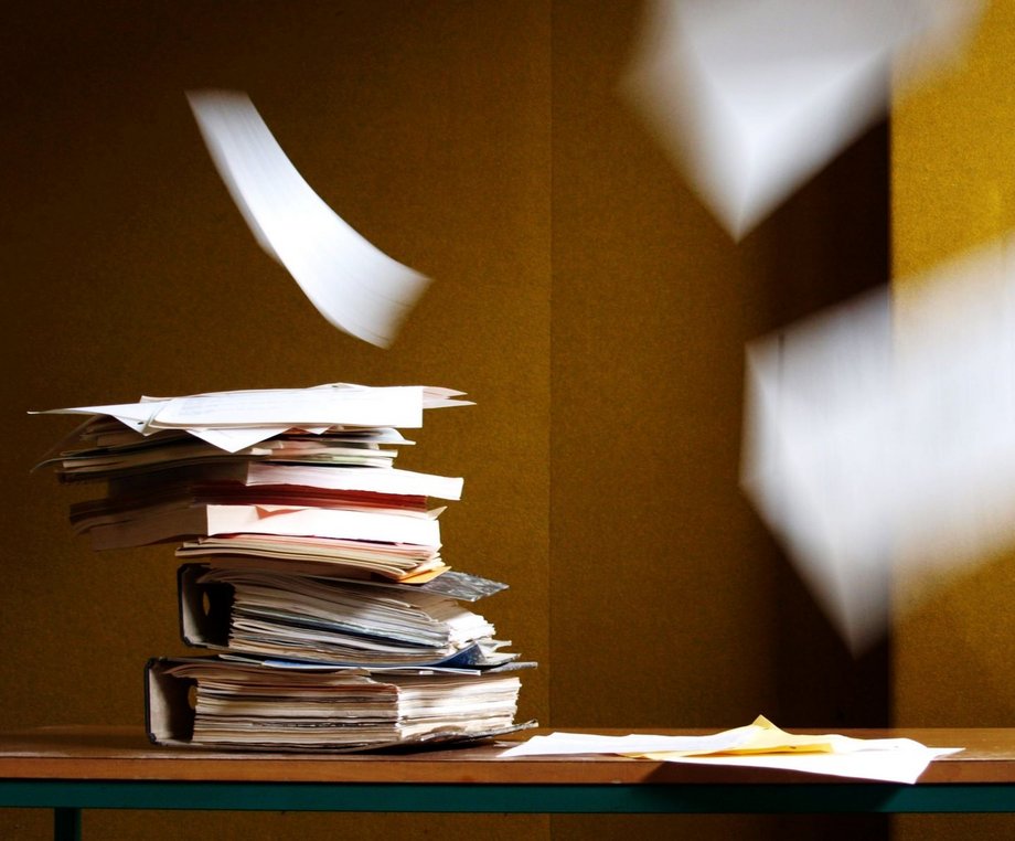 Ein Stapel mit Büchern und Ordnern von welchem einzelne Dokumente durch den Raum geweht werden

