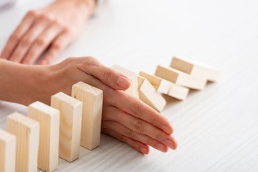 Holzquader stehen in einer Reihe. Die ersten sind umgefallen, eine Hand ist dazwischen, sodass die restliche Reihe stehenbleibt.