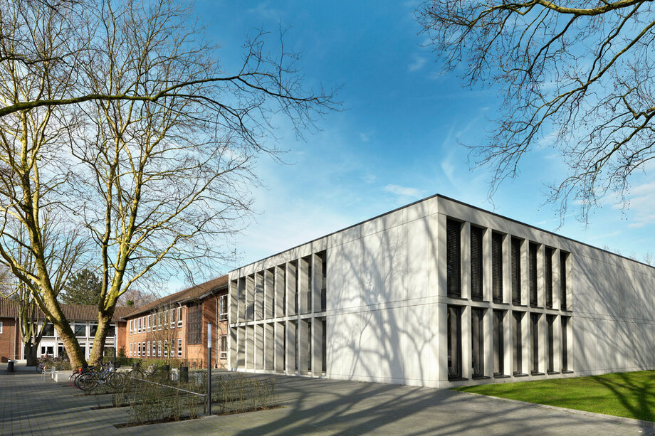 Das Gebäuder der katho in Münster. Ein graues zweistöckiges Gebäude mit einer großen Fensterfront. Rechts im Bild ist ein Schild zu sehen auf dem "KatHO NRW Abteilung Münster" steht.