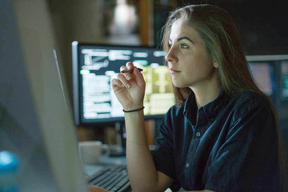 Eine junge Frau sitzt an einem Schreibtisch vor einem Bildschirm. Sie hält einen Stift in der Hand und denkt nach. In dem dunklen Raum ist sie von weiteren Bildschirmen umgeben.