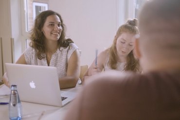 Zwei Studentinnen und ein Student sitzen an einem Tisch mit Laptop.