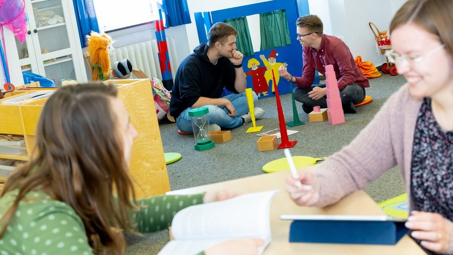 Zwei Studierende sitzen an einem Tisch in einem Spielzimmer, dahinter sitzen zwei Männer auf dem Boden.