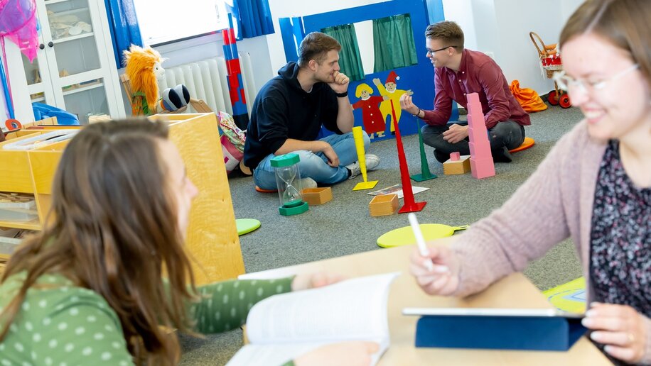 Zwei Studierende sitzen an einem Tisch in einem Spielzimmer, dahinter sitzen zwei Männer auf dem Boden.