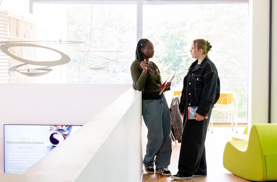 Zwei Studierende unterhalten sich und halten dabei ein Buch und ein Collegeblock.

