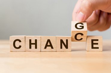 Eine Hand dreht einen Holzwürfel, sodass dessen Aufschrift von C zu G wechselt. Zusammen mit den anderen Holzwürfeln wird aus "Chance" die Aufschrift "Change".