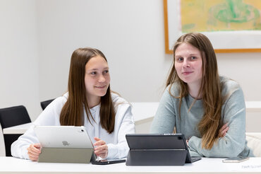 Zwei Studentinnen sitzen während eines Seminars nebeneinander und hören aufmerksam zu.

