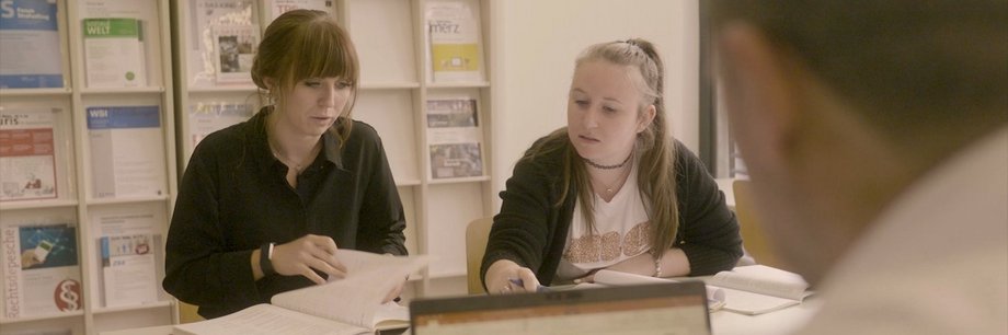Drei Studierende sitzen in der Aachener Bibliothek als Lerngruppe zusammen. Die zwei Studentinnen blättern in Büchern, der Student schaut auf seinen Laptop.