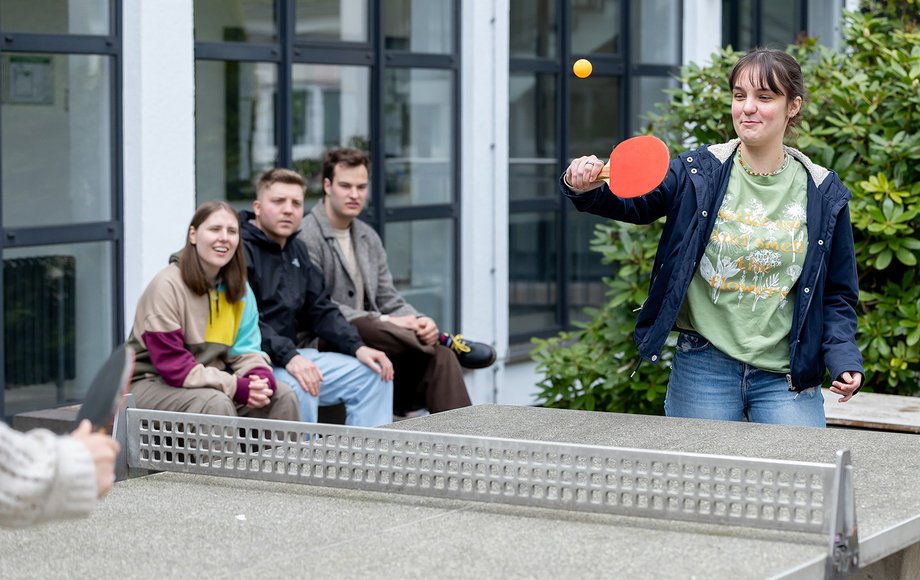 Zwei Studentinnen spielen im Innenhof Tischtennis, drei weitere Studierende gucken Ihnen dabei zu.

