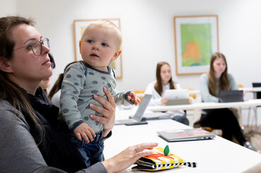 Mehrere Studierende sitzen während eines Seminars nebeneinander und hören aufmerksam zu. Eine Studentin hält ein Baby.

