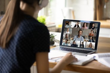 Eine junge Frau sitzt an einem Tisch vor einem aufgeklappten Laptop. Sie trägt Kopfhörer. Auf dem Bildschirm des Laptops sind unterschiedliche Personen zu sehen, die sich alle an einem Online-Meeting beteiligen.