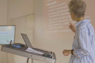 Eine Professorin steht an einem Laptoppult und erklärt etwas zu ihrer Präsentation.