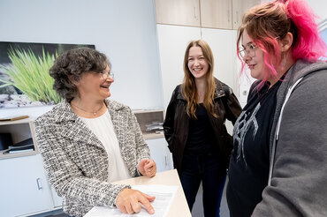 Eine Frau steht in einem Sekretariat und gibt zwei Studierenden einen Zettel. 