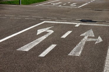 Auf dem Asphalt sind mehrere weiße Pfeilmarkierungen zu sehen, die verschiedene Fahrtrichtungen (links, geradeaus und rechts) anzeigen.