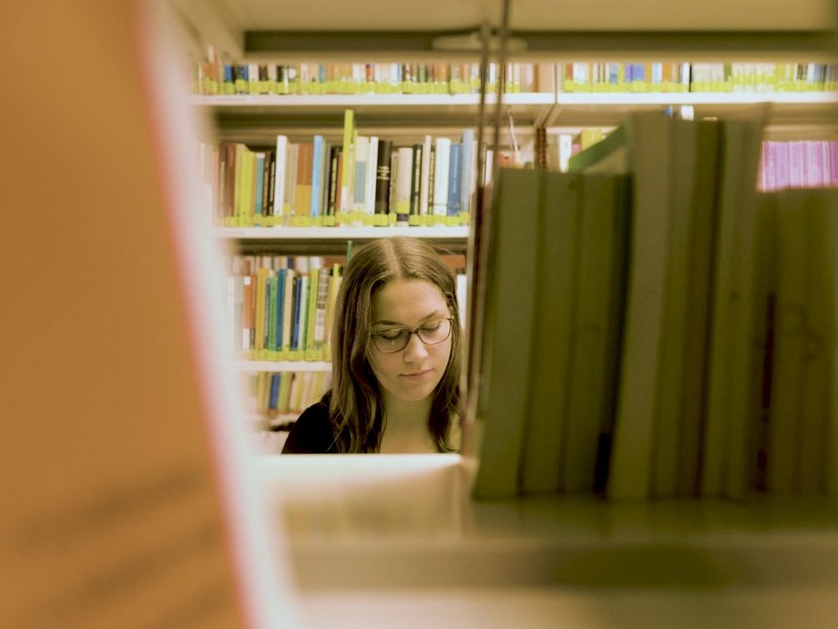 Blick durch ein Bibliotheksregal auf eine Studentin.